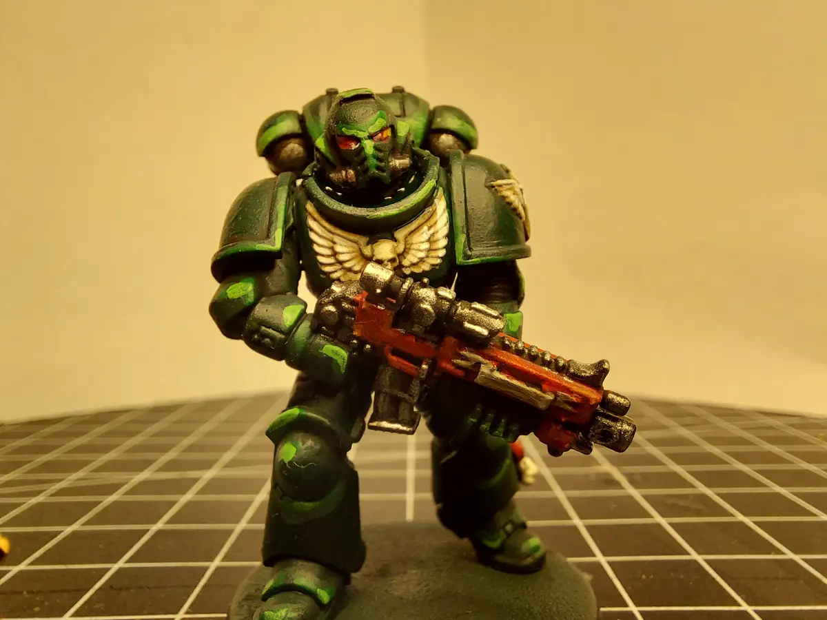A painted miniature of a green-armored Space Marine with glowing red eyes and a red weapon stands on a grid-patterned…