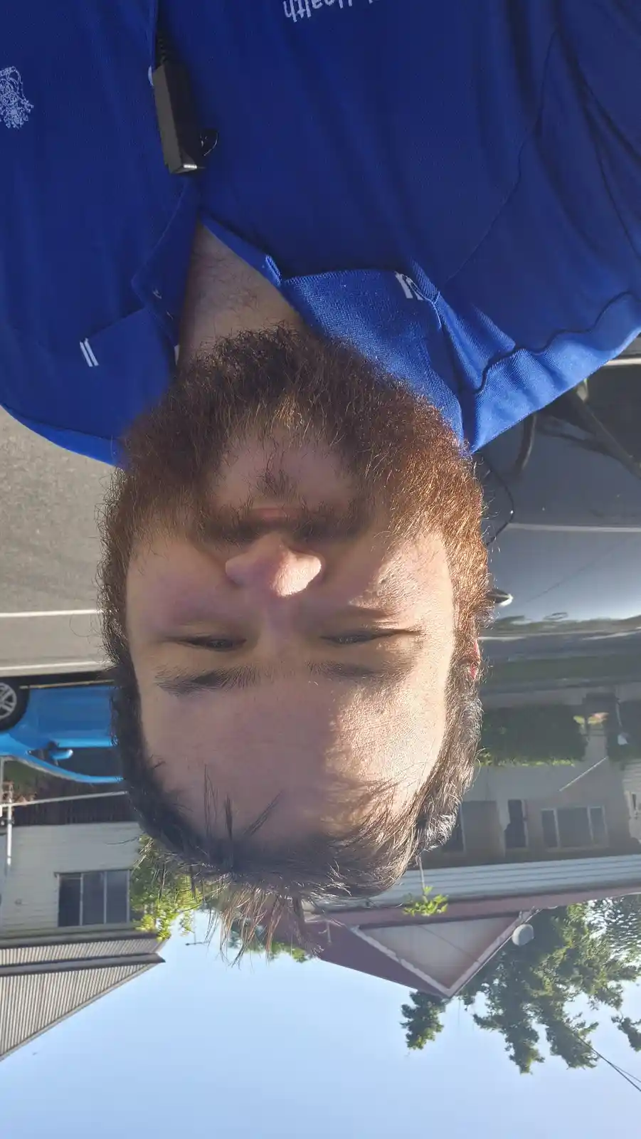 A vertically flipped selfie shows a man in a blue polo shirt with a lavalier mic clipped to his collar, looking down with…