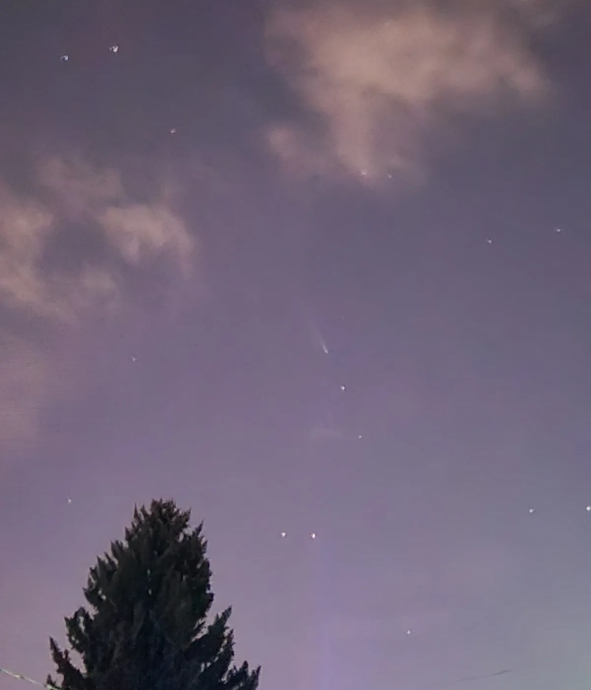 A night sky with scattered stars and faint clouds, featuring a bright streak resembling a comet or meteor. The top of a dark…
