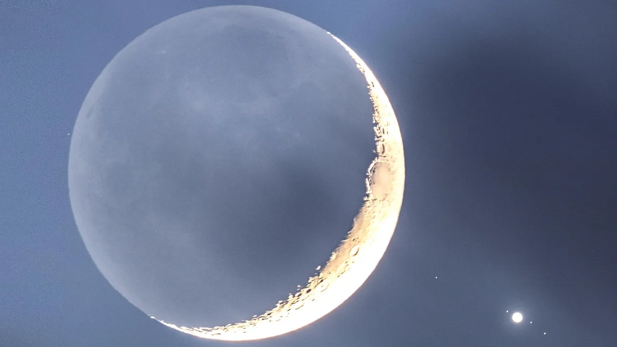 A crescent moon is visible in a dark blue sky, with its illuminated edge showing craters and texture. A small, bright star…