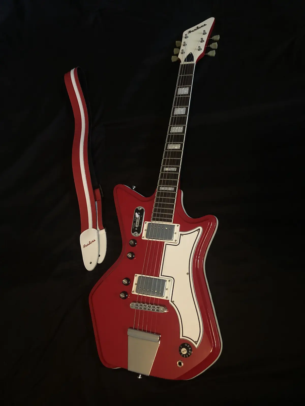 A red and white Airline electric guitar with a matching red and white strap lies on a black fabric background. The guitar…