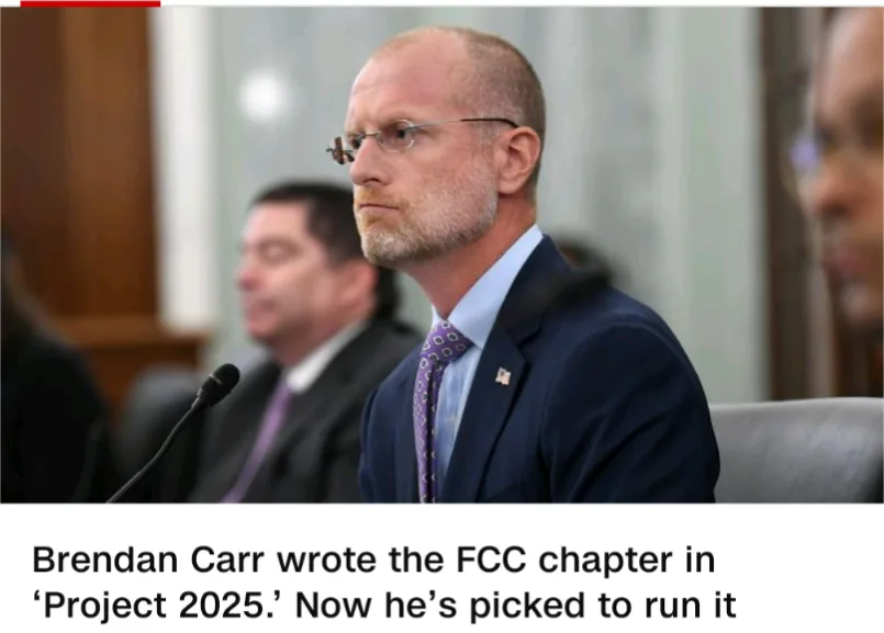 A man in a dark suit and glasses, identified as Brendan Carr, sits at a hearing with a microphone in front of him. The…