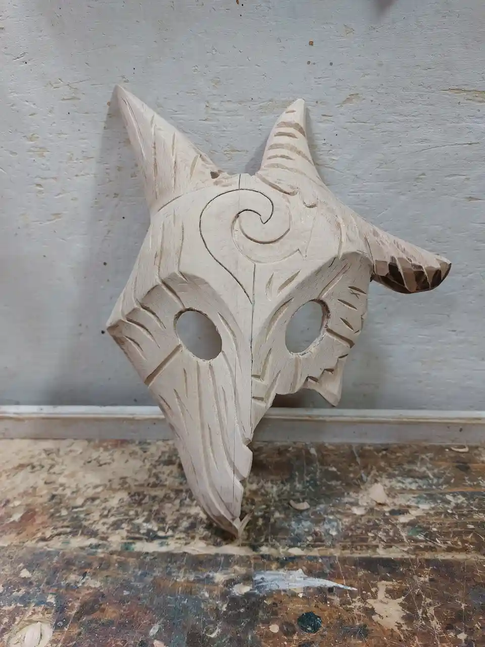 A wooden, hand-carved mask resembling a demonic or fantasy creature with pointed horns, hollow eye sockets, and intricate…