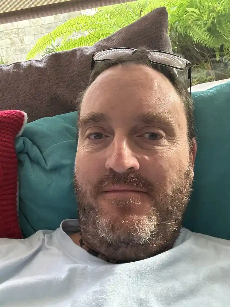 A man with a beard and glasses resting on his head lies on a couch surrounded by pillows, looking directly at the camera…
