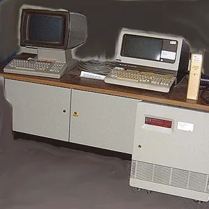 Two vintage beige Apple Macintosh computers sit side-by-side on a wooden desk with a gray cabinet underneath. The computer…