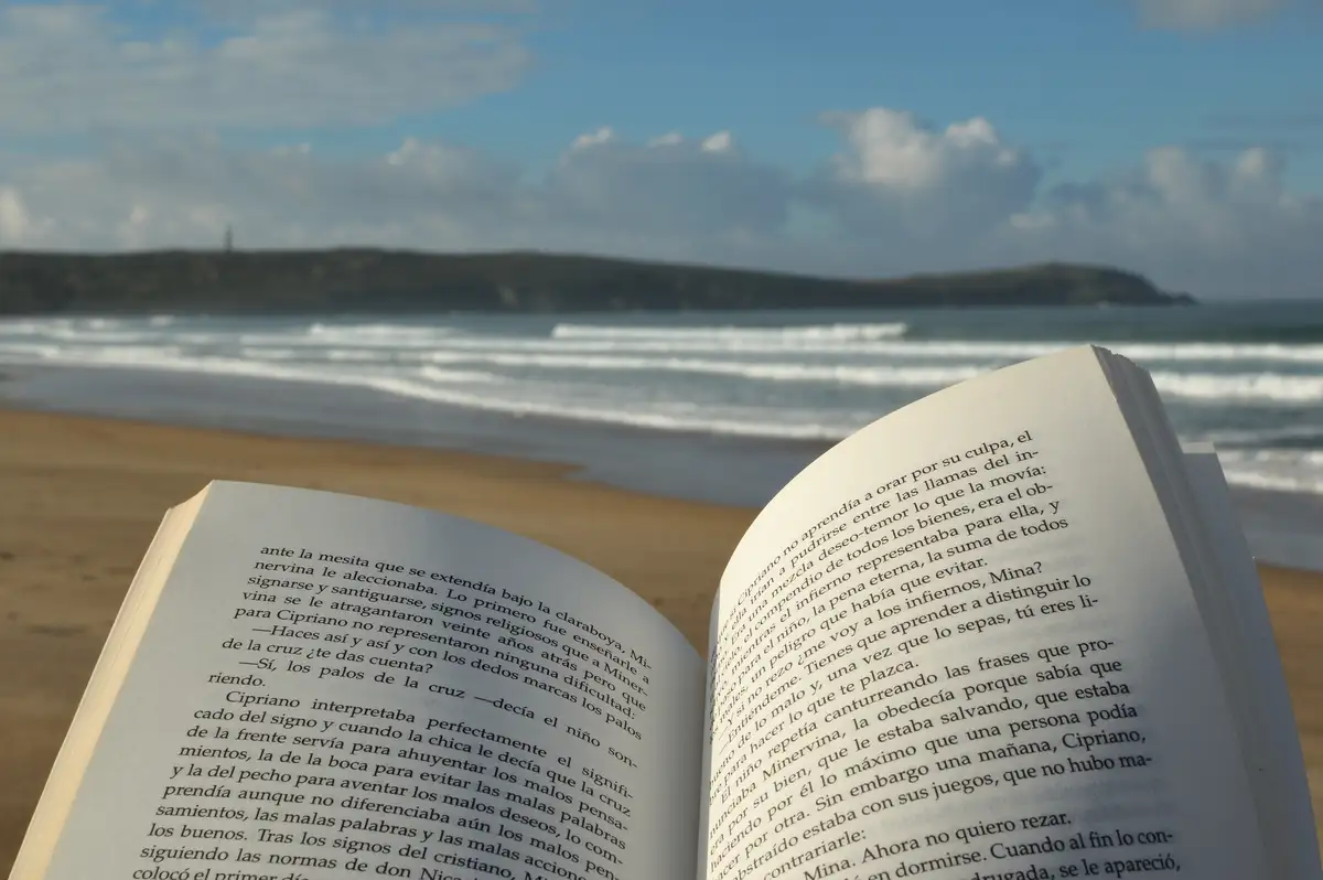 An open book with Spanish text lies on a sandy beach, with ocean waves and a distant headland under a partly cloudy sky. The…