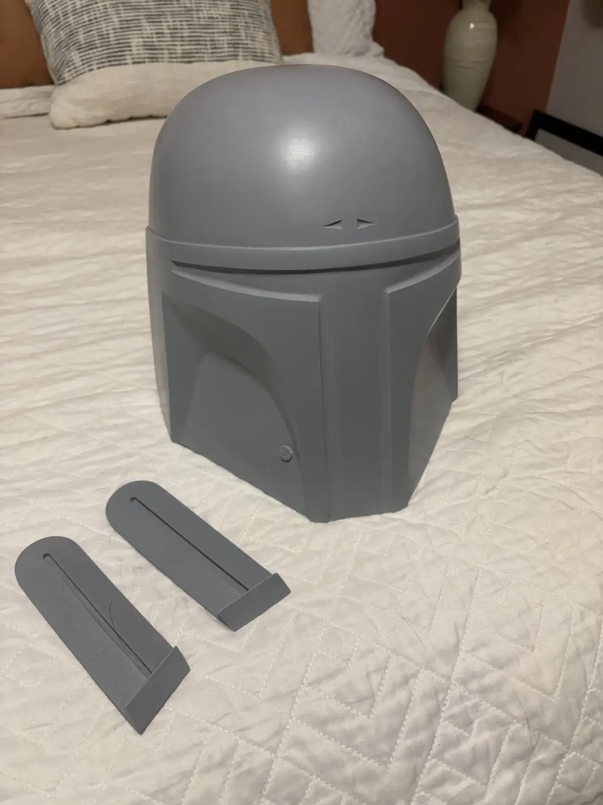 A gray-painted Boba Fett helmet sits on a white quilted bed, with two gray, detached, rectangular pieces resembling the…