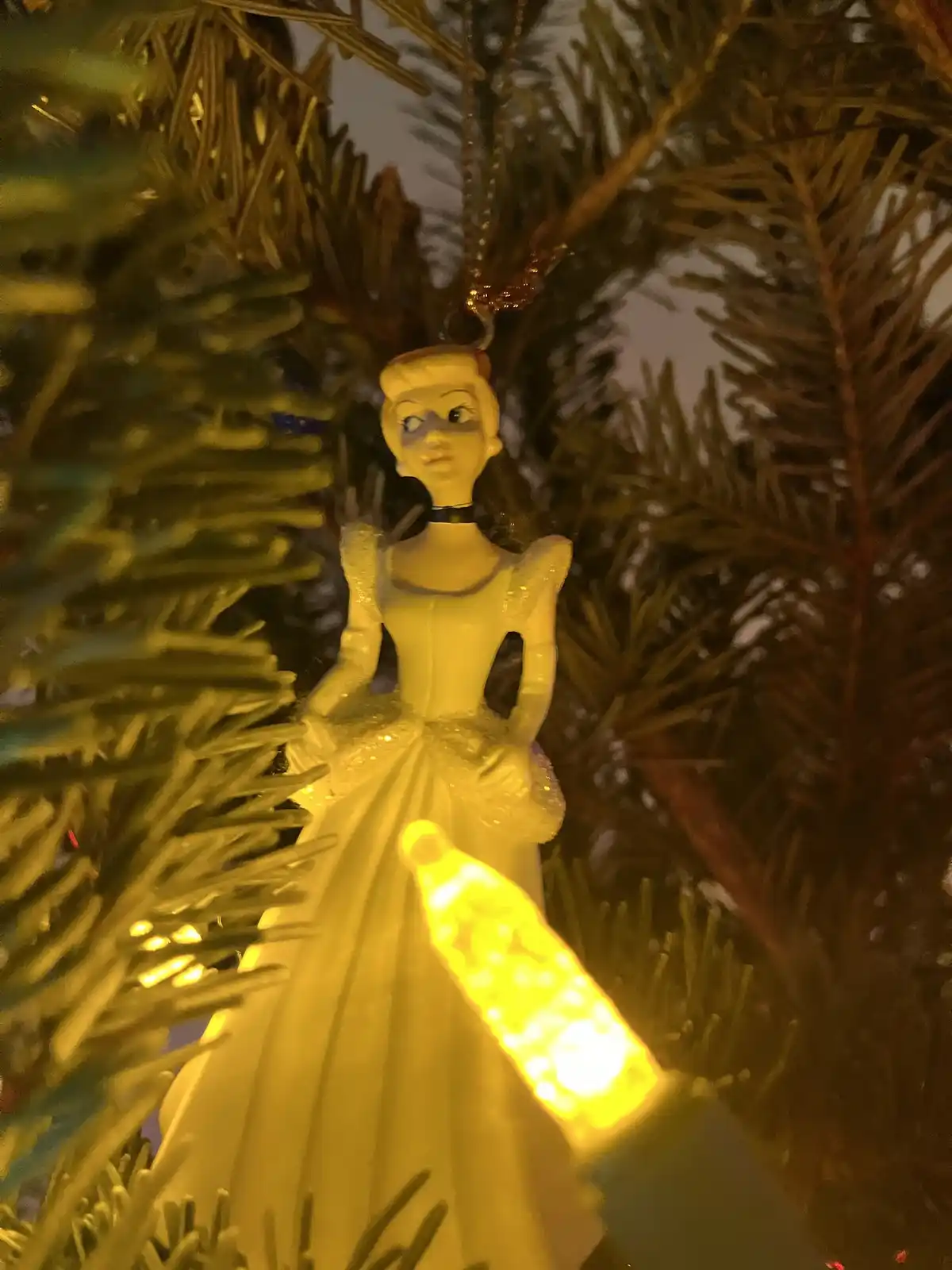 A close-up photo of a Cinderella Christmas ornament hanging on a tree, illuminated by a warm yellow string light that casts…