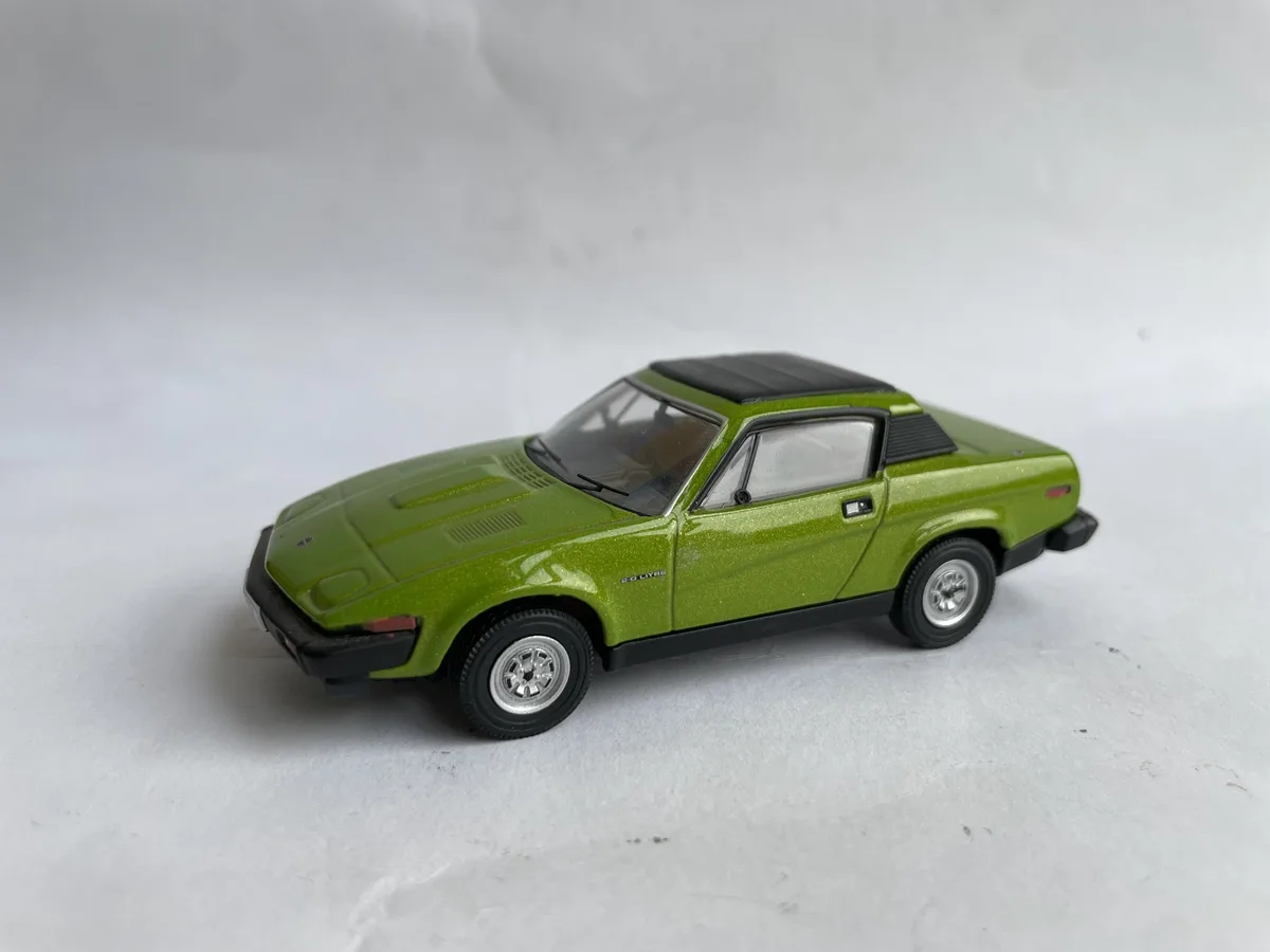 A green die-cast model of a classic sports car, likely a Triumph TR7, is photographed against a plain white background. The…