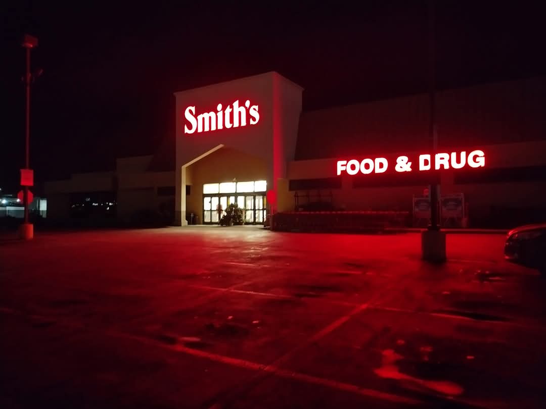 A nighttime photo of a Smith's Food & Drug store with its red neon signs glowing brightly against the dark sky. The parking…