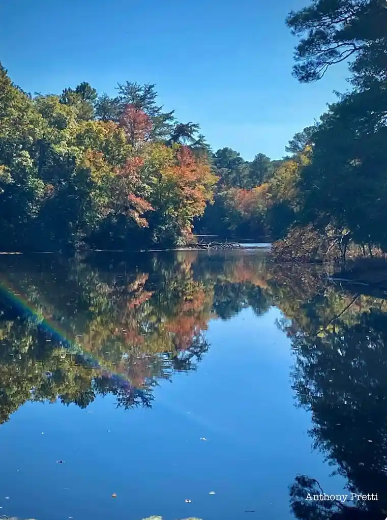 A serene autumn landscape featuring a calm river reflecting colorful trees with orange, red, and green foliage under a clear…