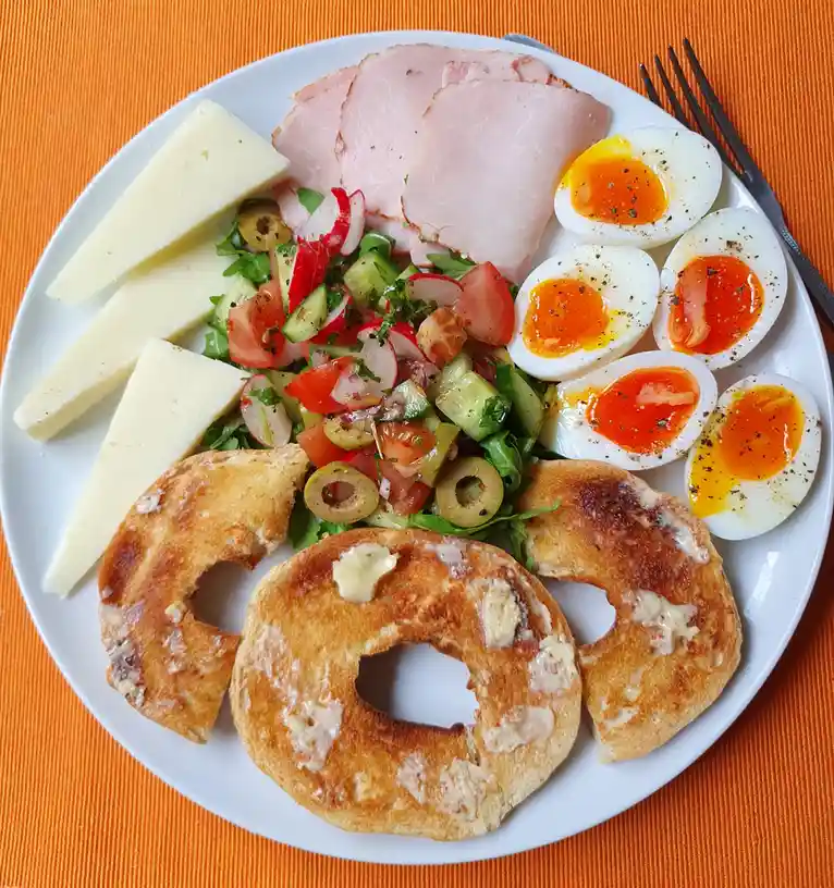 A white plate holds a breakfast spread including sliced ham, hard-boiled eggs cut in half, cheese wedges, a fresh salad with…