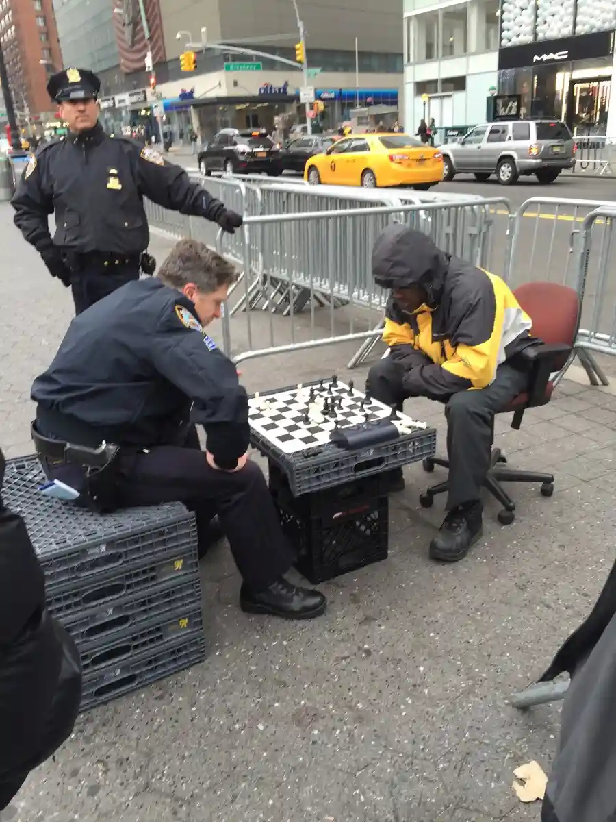Two police officers are playing chess on a street with a man in a yellow and black jacket. One officer is seated on crates…