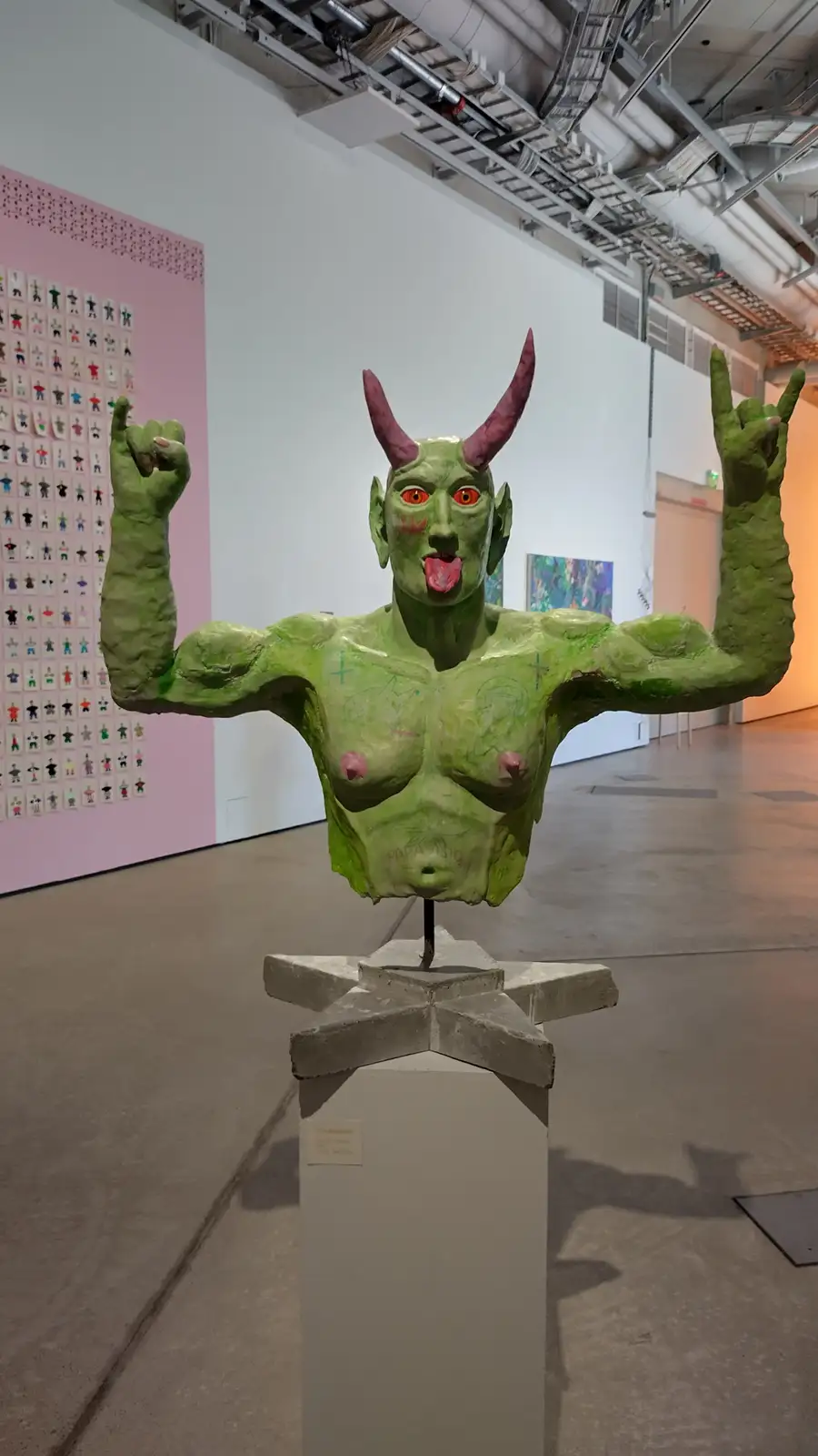A green, muscular, horned sculpture of a devil-like figure with red eyes and tongue sticking out, standing on a pedestal in…
