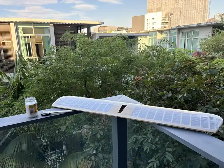 A homemade solar panel, constructed from wood and covered in solar cells, rests on a balcony railing next to a jar of white…