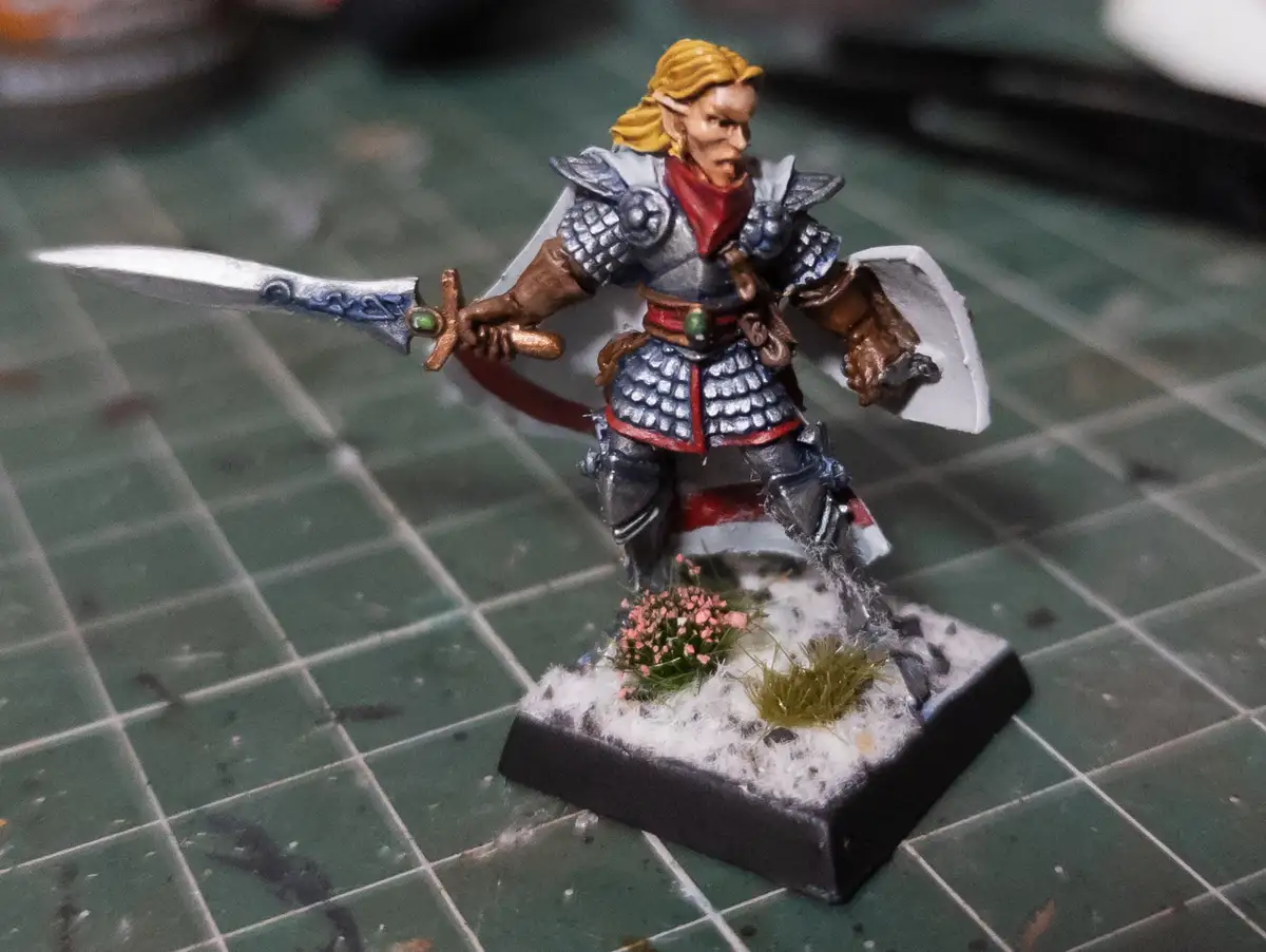 A painted fantasy miniature of an elf warrior with blonde hair, holding a sword in one hand and a shield in the other…
