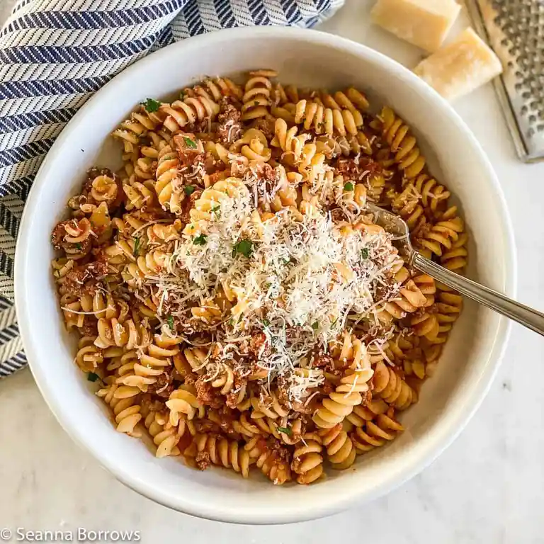 A top-down photo of a white bowl filled with rotini pasta in a meat sauce, topped with grated cheese and chopped parsley. A…