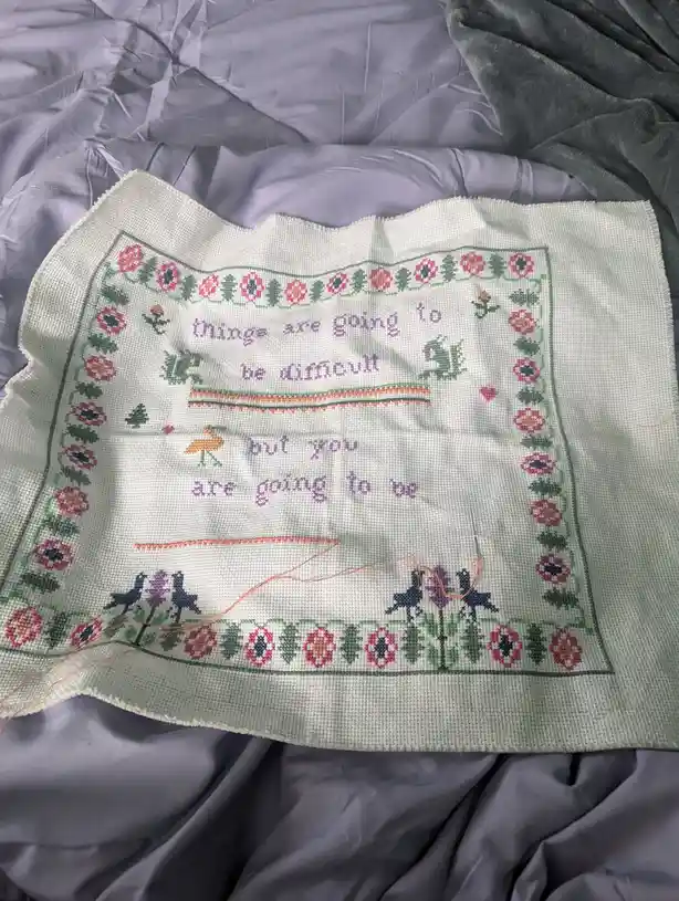 A partially completed cross-stitch piece on white fabric, featuring a floral border and small stitched birds, lies on a grey…