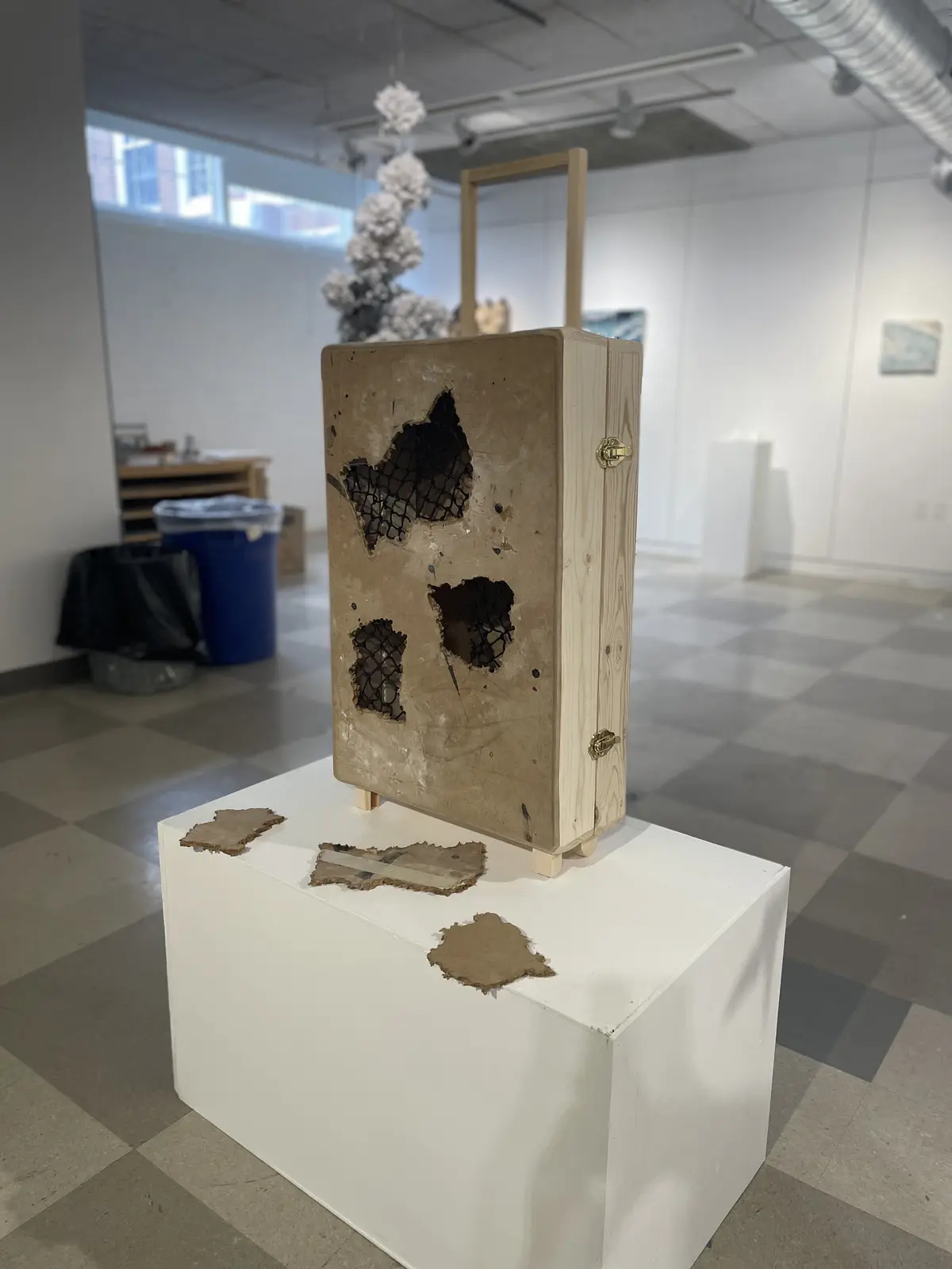 A wooden box sculpture with large torn holes revealing black netting inside, displayed on a white pedestal in an art…