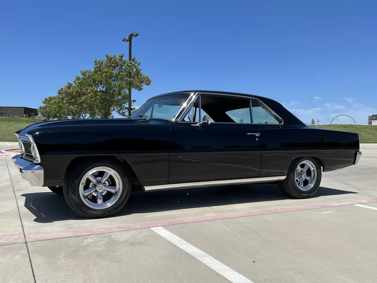 A shiny black classic American muscle car, likely a Chevrolet, is parked in a sunny parking lot with a clear blue sky. The…