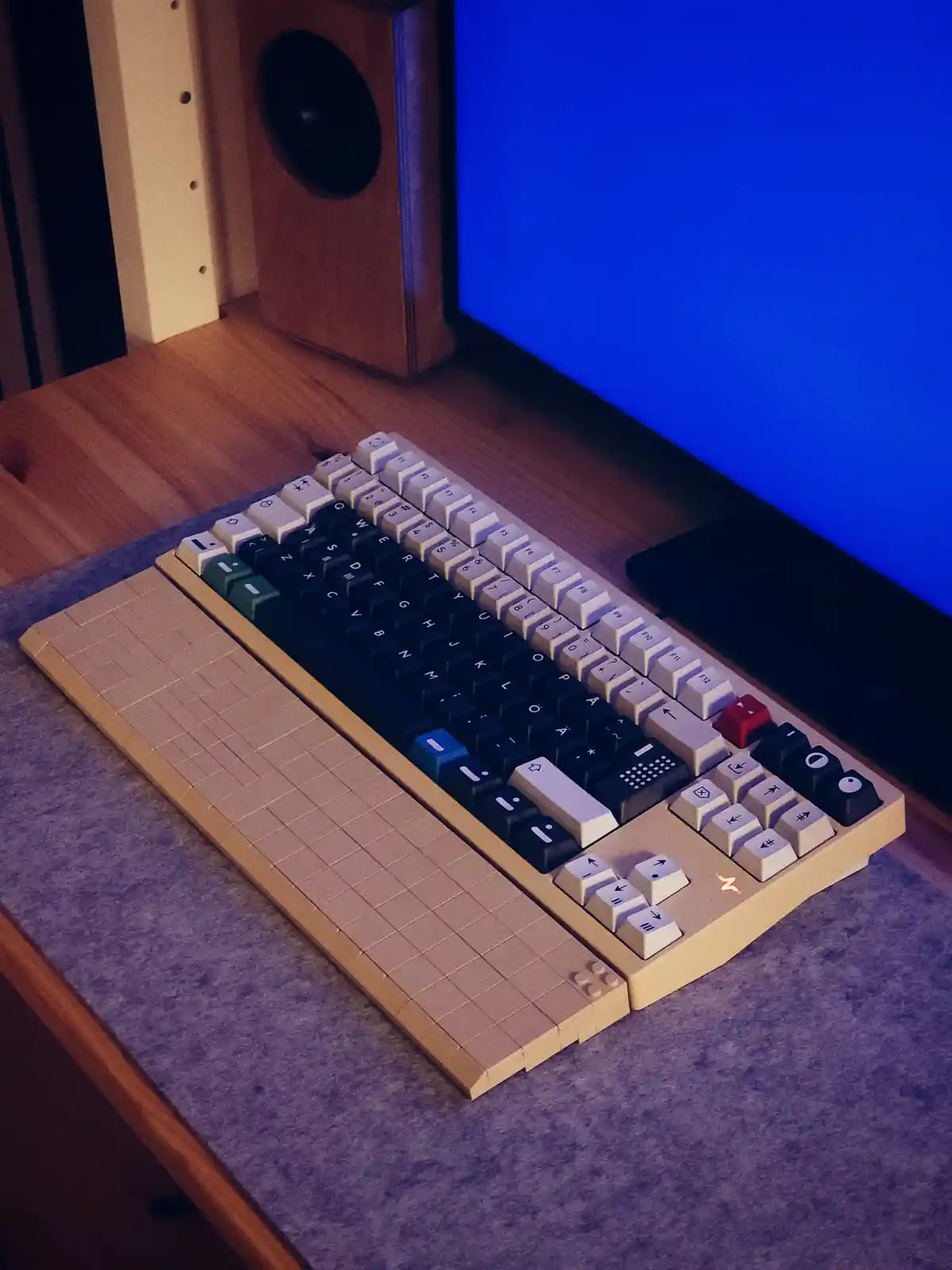 A custom-built keyboard with a tan, grid-patterned base resembling LEGO bricks sits on a desk next to a blue monitor and…