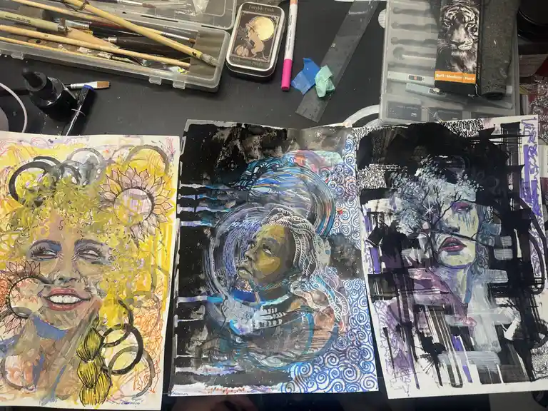 Three distinct, colorful, and stylized portrait artworks are displayed side-by-side on a cluttered artist's desk, surrounded…