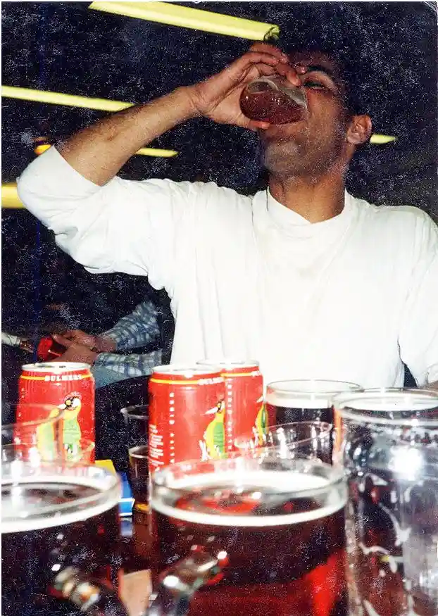 A man in a white shirt is drinking from a glass while surrounded by numerous beer cans and glasses on a table. The overlaid…