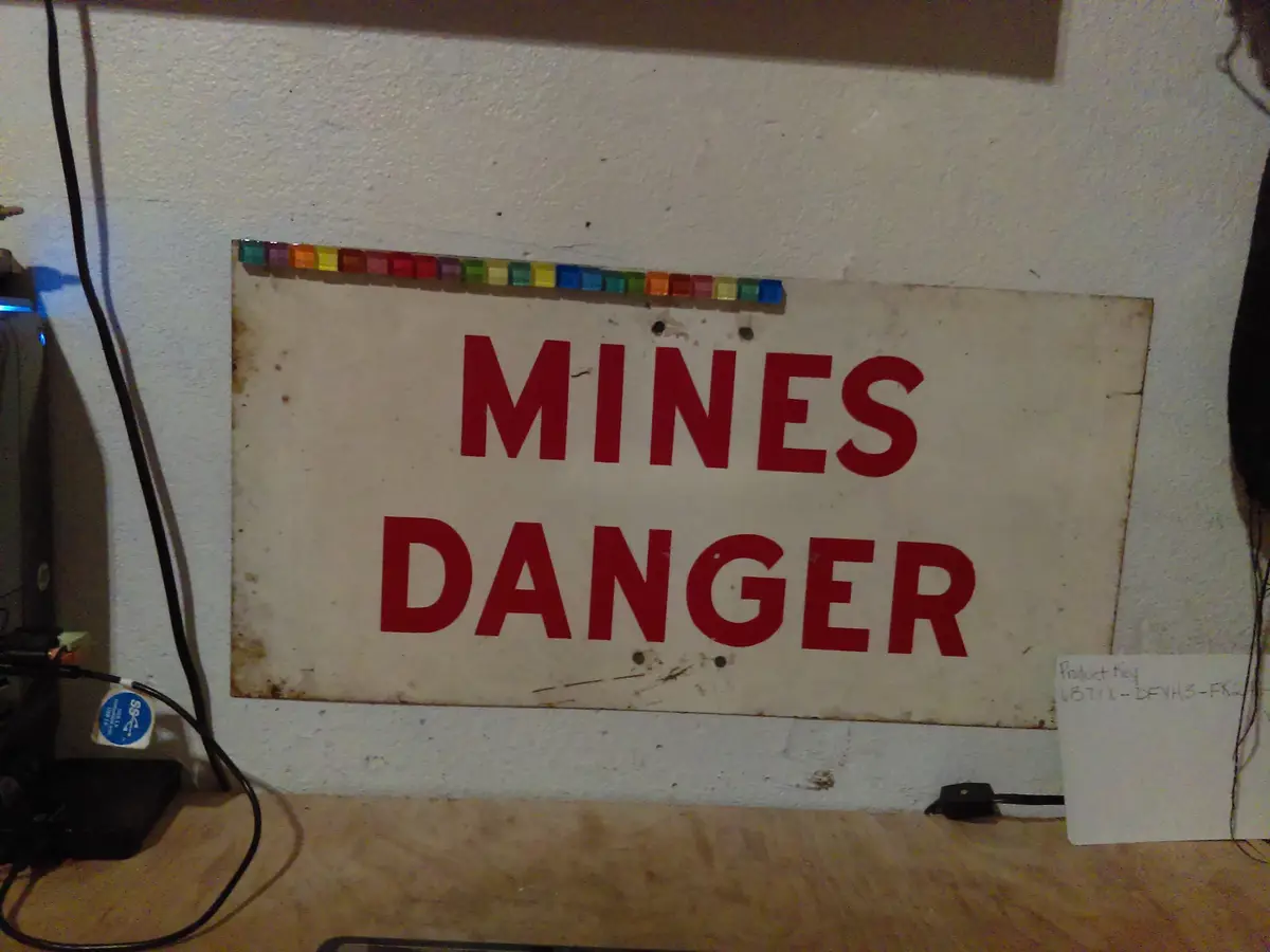 A weathered white sign with bold red text reading 'MINES DANGER' is mounted on a wall, with a row of colorful translucent…