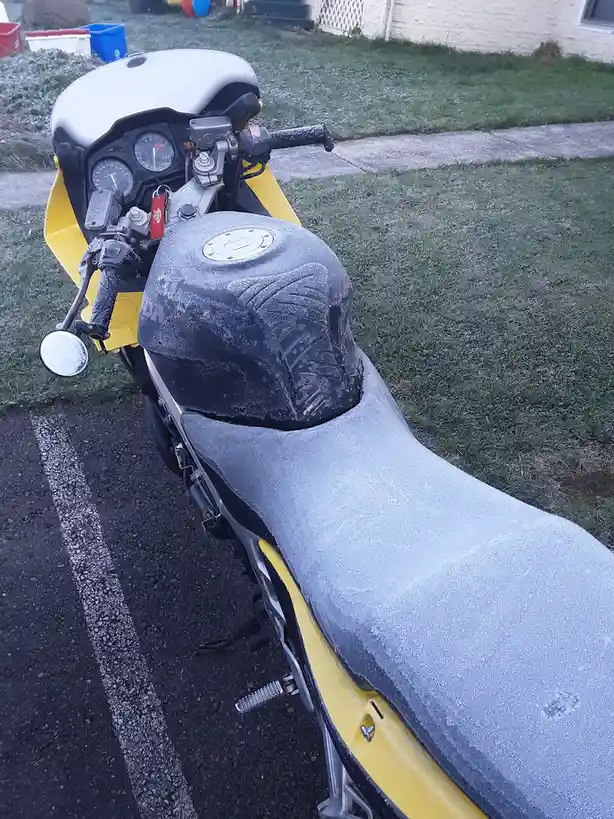 A yellow motorcycle is parked on a driveway with its seat, fuel tank, and handlebars covered in a thick layer of frost…