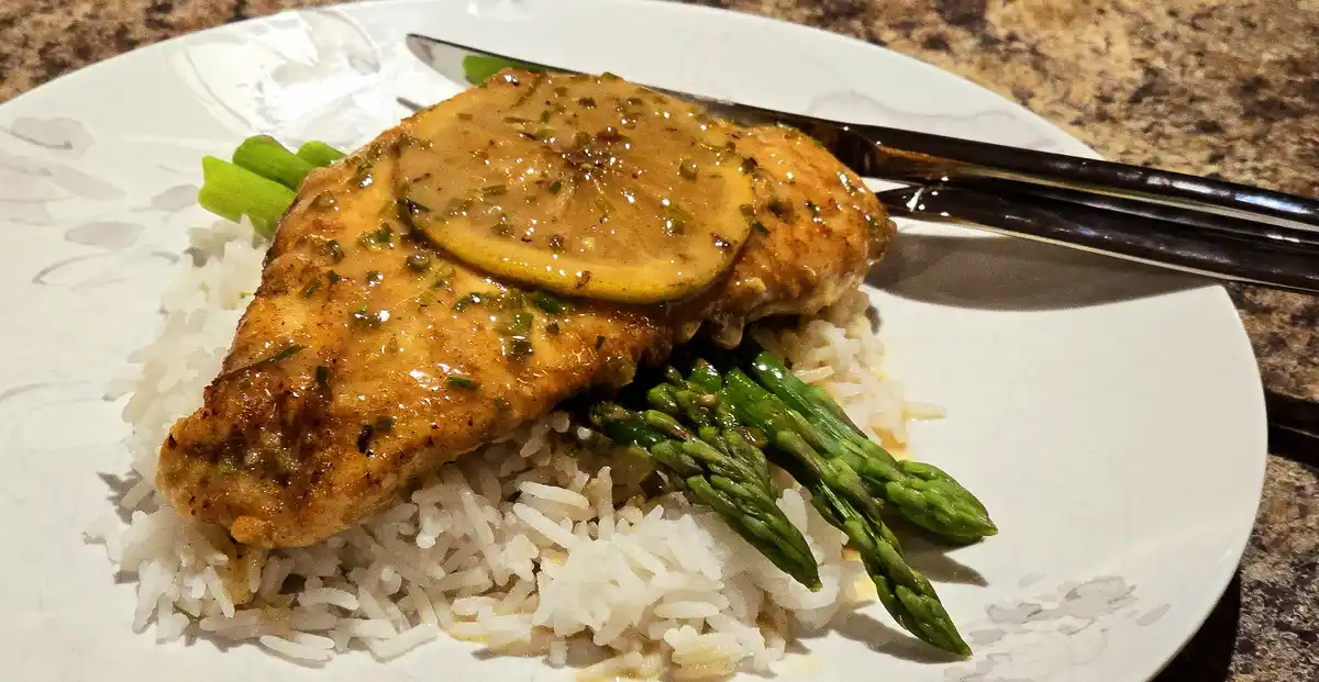 A plated meal featuring a golden-brown chicken breast topped with a lemon slice and herbs, served over white rice with green…