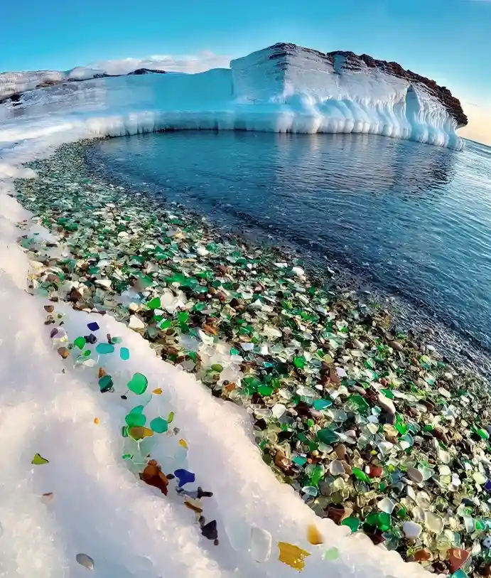 A stunning natural landscape shows a shoreline covered in colorful, fragmented glass pieces, nestled between snow and a calm…