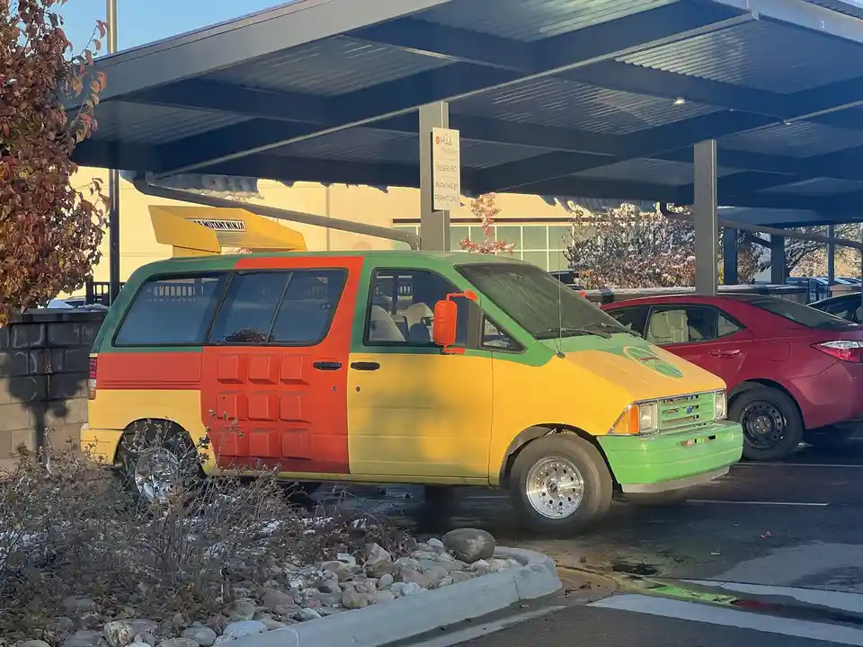 A brightly colored van parked in a covered lot, painted in sections of green, red, and yellow with a textured red panel on…