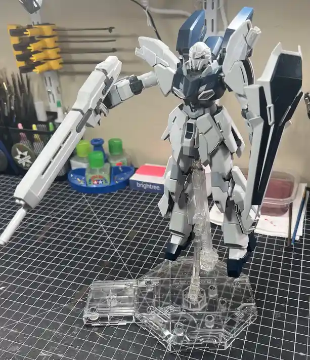 A white and blue Gundam-style model kit stands on a clear plastic display stand on a black grid-patterned work mat, with its…