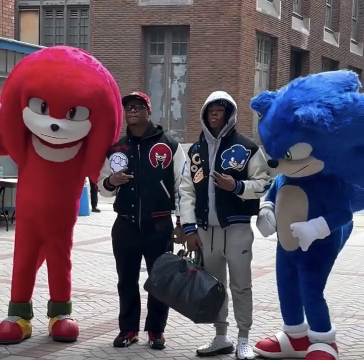 Two men in Sonic the Hedgehog-themed varsity jackets stand between full-body costumes of Knuckles the Echidna and Sonic the…