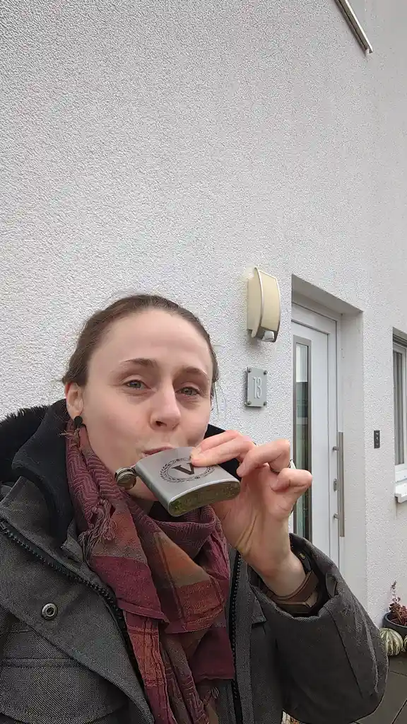 A person stands outside a house with the number 13 on the wall, drinking directly from a silver flask with a 'V' logo. They…