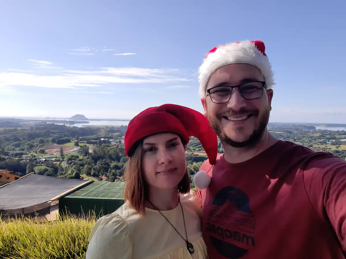 A selfie of a smiling man and woman on a hilltop overlooking a green landscape and water, both wearing red Santa hats. The…