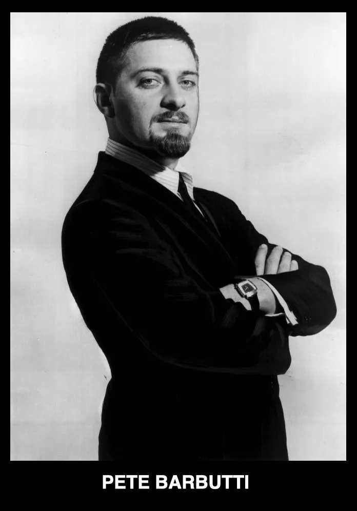 A black-and-white portrait of a man with short dark hair and a goatee, wearing a dark suit and striped shirt, standing with…