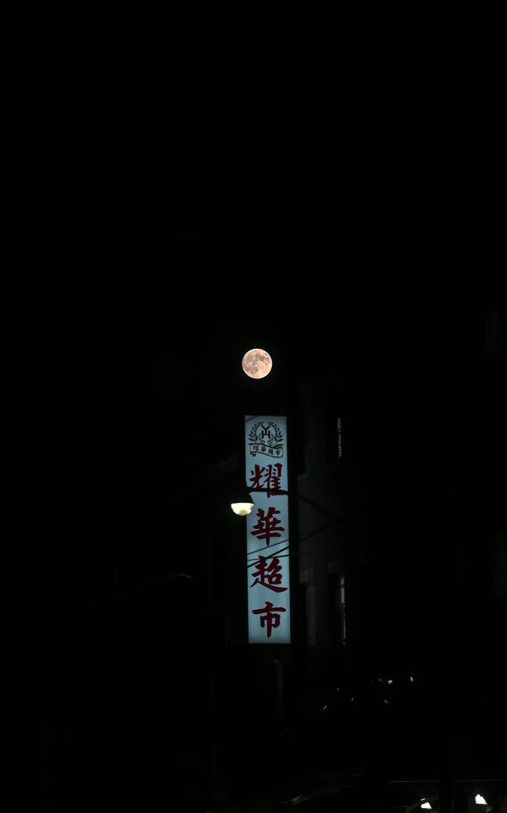 A full moon shines brightly in a dark night sky, positioned directly above a vertically oriented, illuminated sign for a…