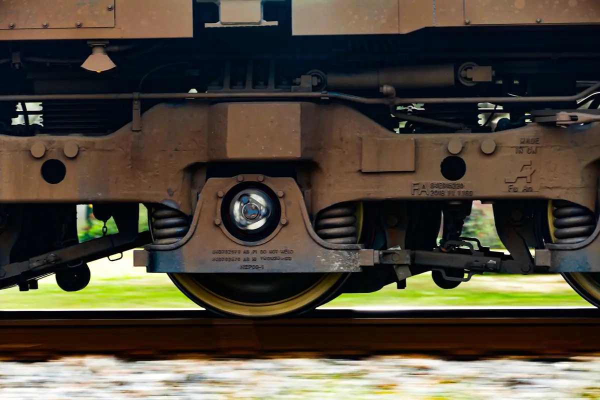 A close-up photo of a moving train's undercarriage, showing rusted metal components, wheels, and suspension springs. The…