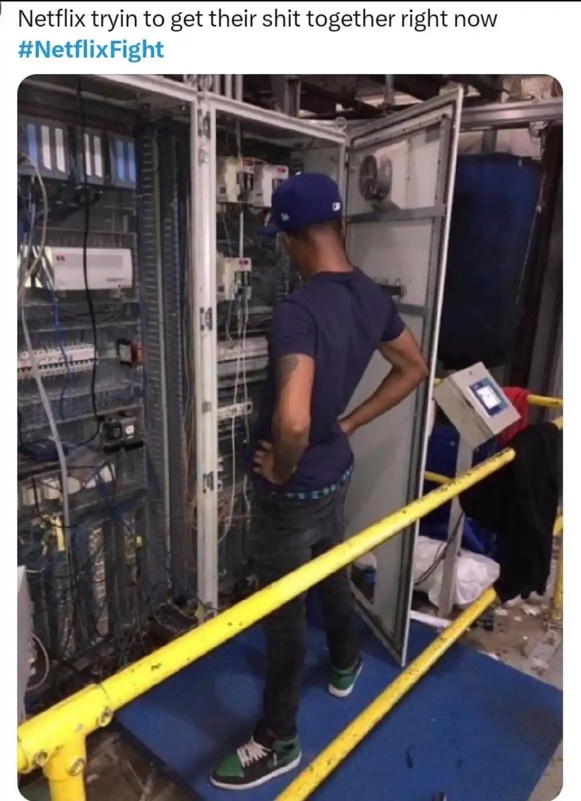 A man in a blue cap and dark t-shirt stands with hands on hips, facing open server racks filled with tangled wires and…