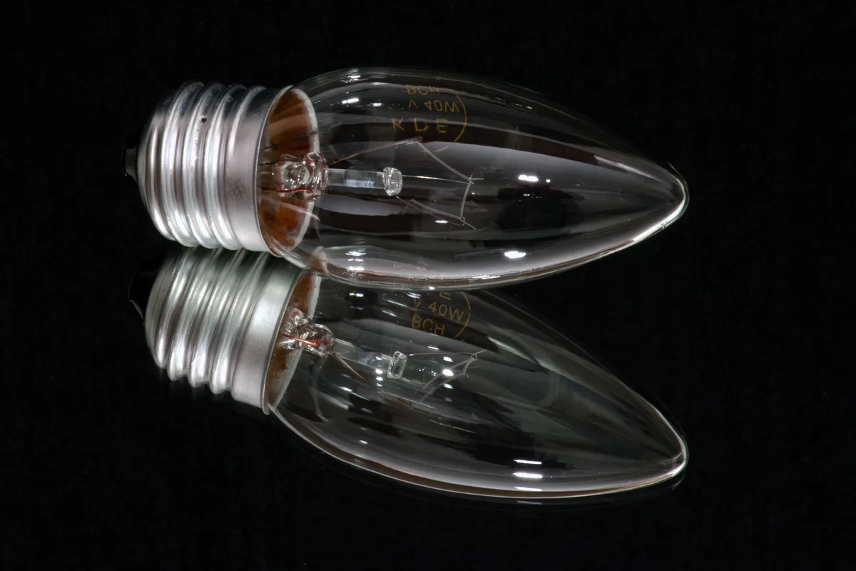 A clear, unlit candle-style incandescent light bulb with a metal screw base rests on a reflective black surface, creating a…