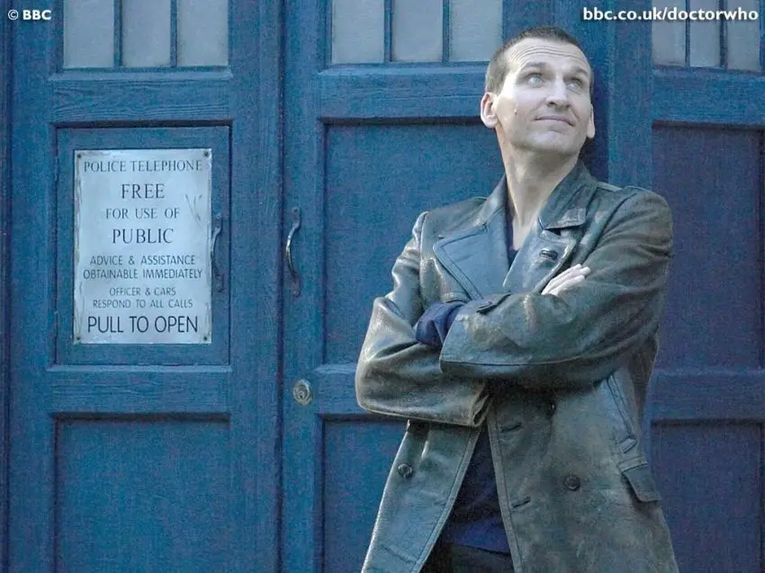 A man in a worn leather coat stands with arms crossed in front of a blue police box door, looking upward with a smug…