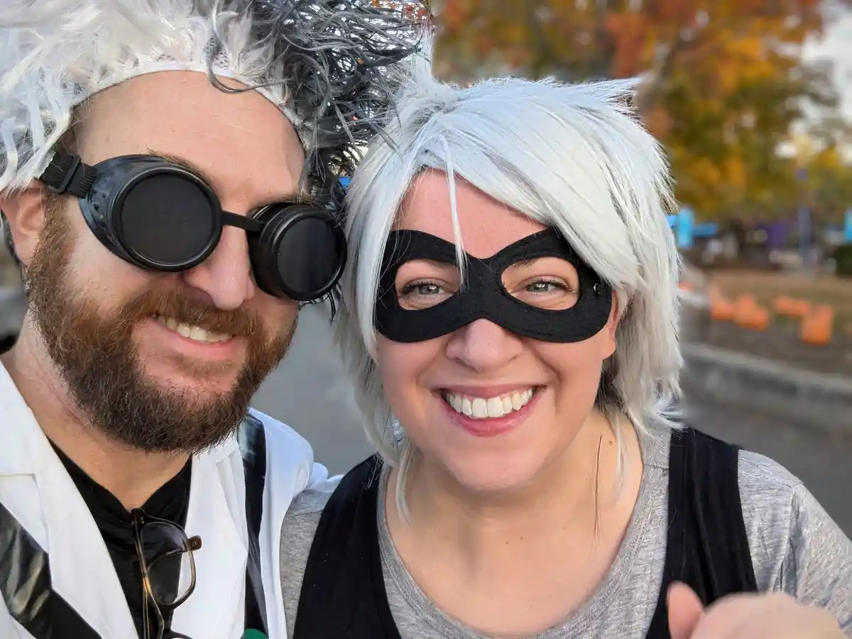 Two people in Halloween costumes pose for a selfie; the man on the left wears a white lab coat, a wild white-and-gray wig…