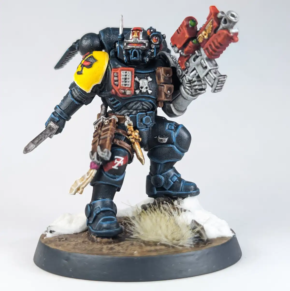 A painted miniature of a heavily armored, futuristic warrior in blue and black armor with red and yellow accents, holding a…