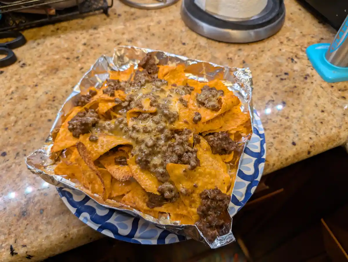 A close-up photo shows a foil-lined paper plate filled with nachos topped with melted cheese and ground beef, sitting on a…