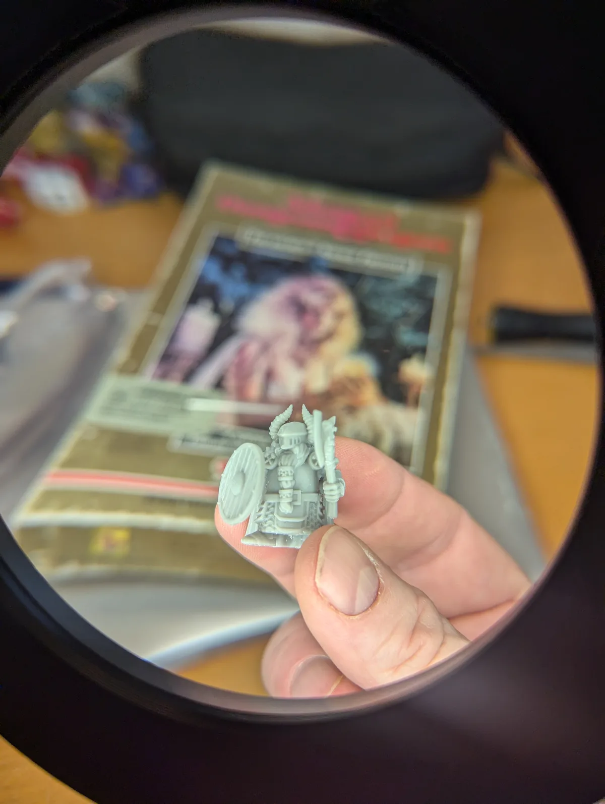 A close-up view through a magnifying glass shows a person's fingers holding a small, unpainted grey miniature of a fantasy…