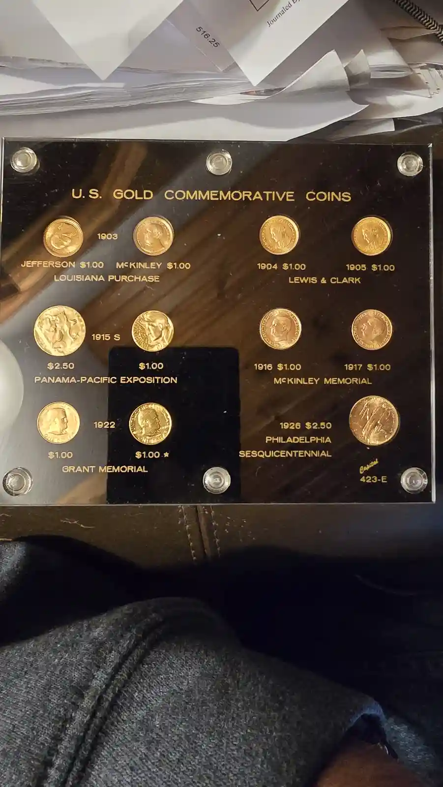 A display case holds a collection of U.S. Gold Commemorative Coins from 1903 to 1926, each labeled with its year…