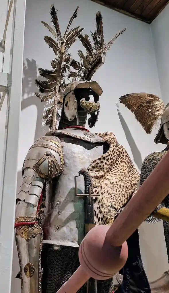 A museum display of ornate medieval armor with a helmet topped by large feather plumes and a leopard-print fur draped over…