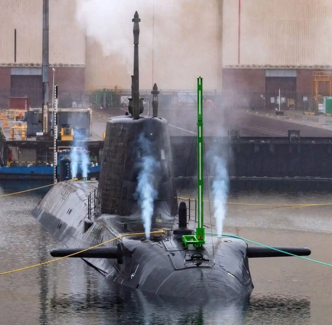 A black submarine is docked in a shipyard, emitting blue steam from multiple vents on its deck. A green crane-like structure…