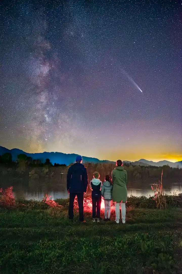 A family of four stands with their backs to the camera, gazing at a breathtaking night sky filled with stars and the Milky…